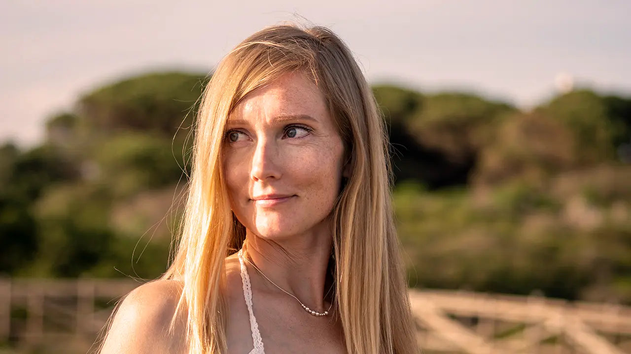 Pitkähiuksinen nainen dyynirannalla kaulassaan hopeinen kaulakoru, jossa lukee morseaakkosilla kirjoitettuna Upea.